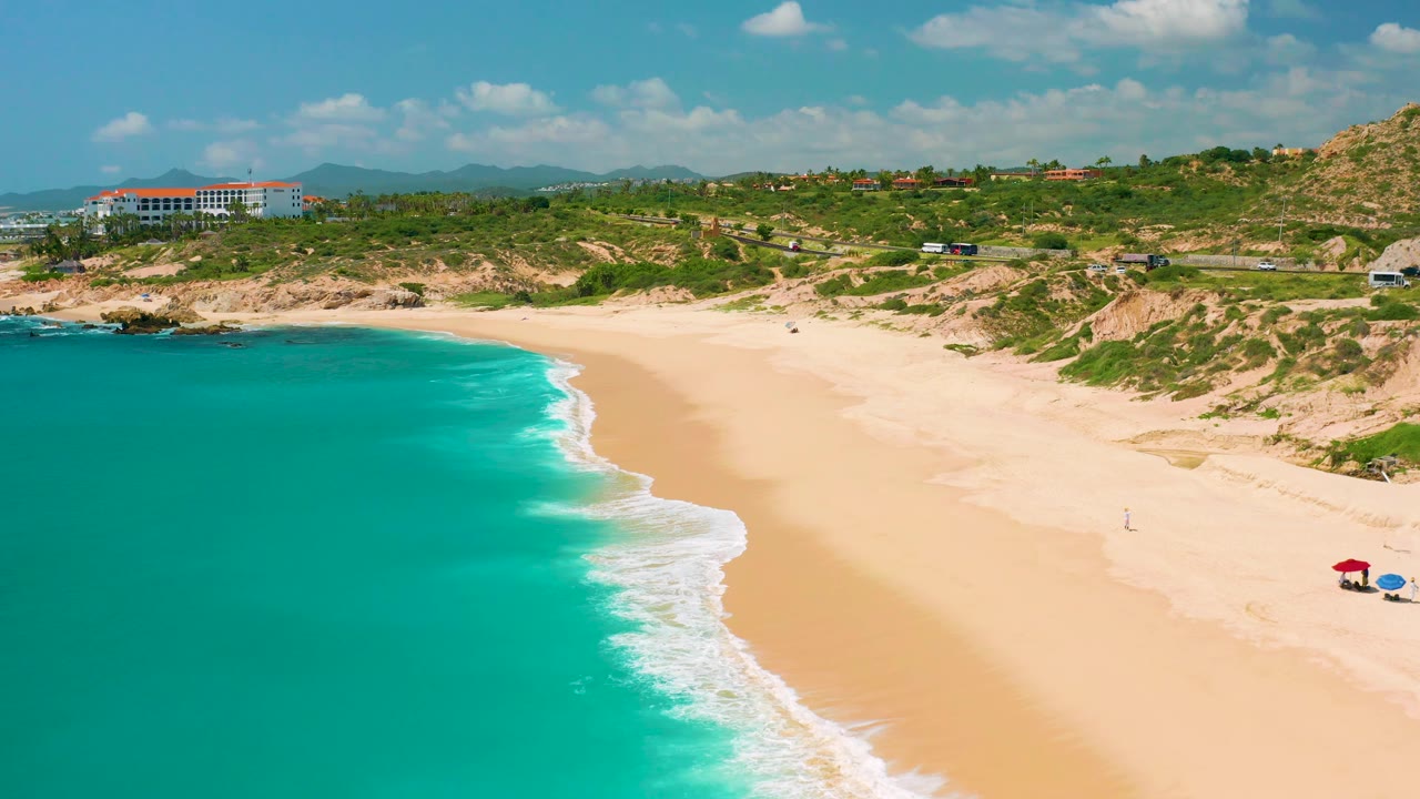 Dreams Beach aerial frame with the ocean filling the left side, a curved white surf band, broad yellow sand, scrub-covered dunes, and small umbrellas near the lower right edge.