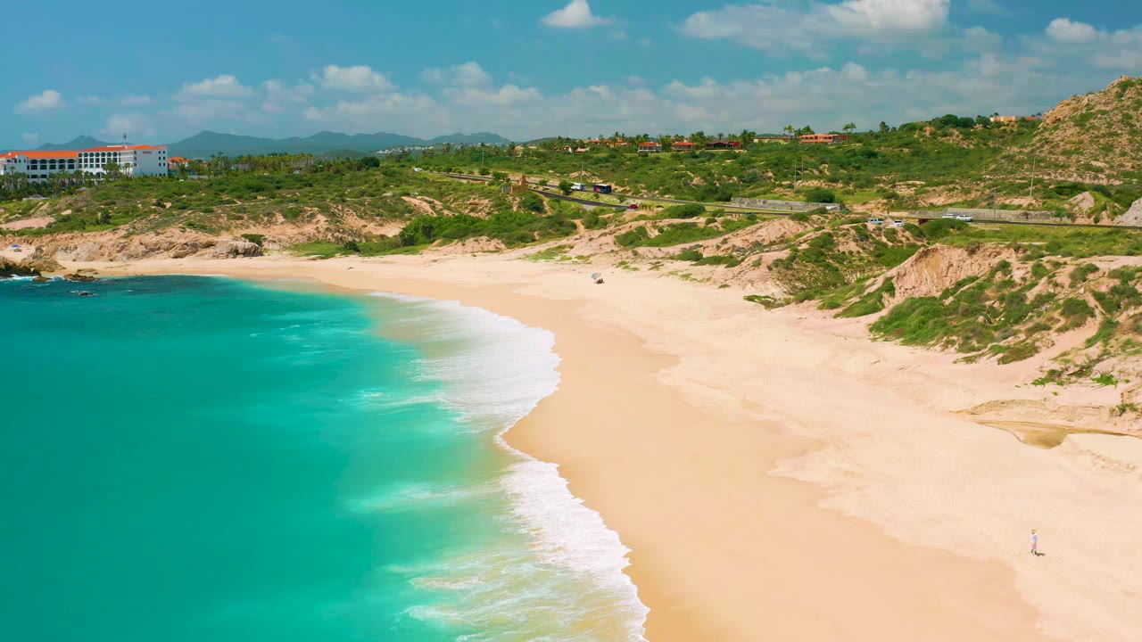 Second aerial frame over Dreams Beach in Baja California Sur, Mexico, showing clear turquoise water curving into pale sand, dune vegetation, a road line, distant hillside buildings, and no visible people.
