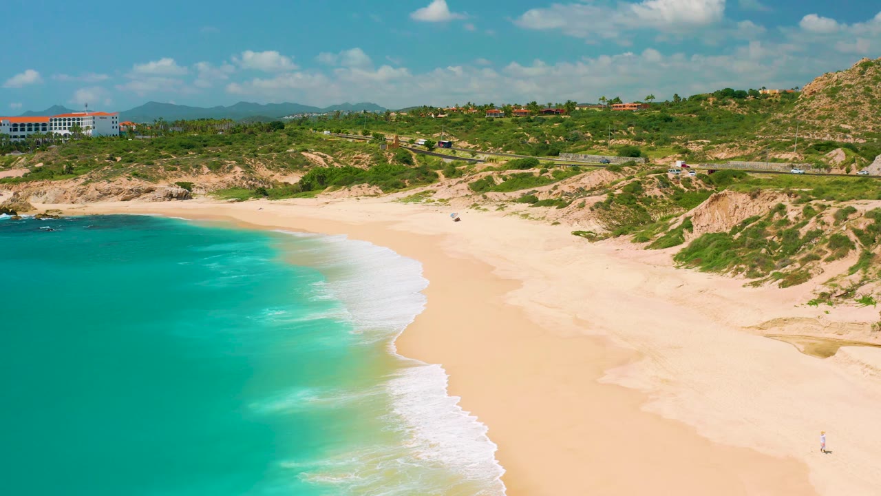 A high aerial frame of Dreams Beach in Baja California Sur, Mexico, where a bright turquoise water band and white surf run along a wide empty beach beneath dunes, road, hillside greenery, and no visible people.