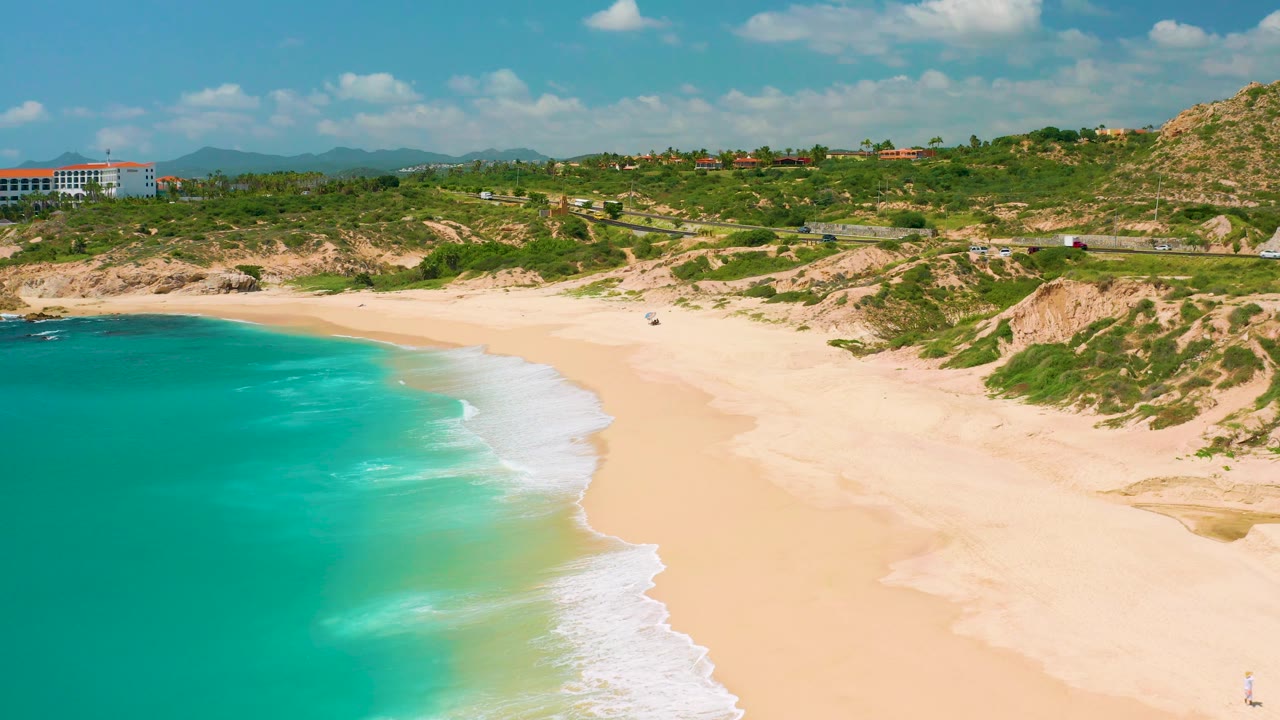 Aerial frame at Dreams Beach in Baja California Sur, Mexico, with turquoise water at the left edge, white surf on empty sand, dune vegetation, a visible road, distant buildings, and no visible people.