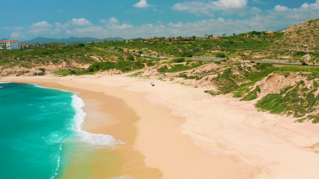 Aerial view over Dreams Beach in Baja California Sur, with turquoise water at left, pale sand across the center, green hillside beyond, a coastal road, distant mountains, and no visible people.