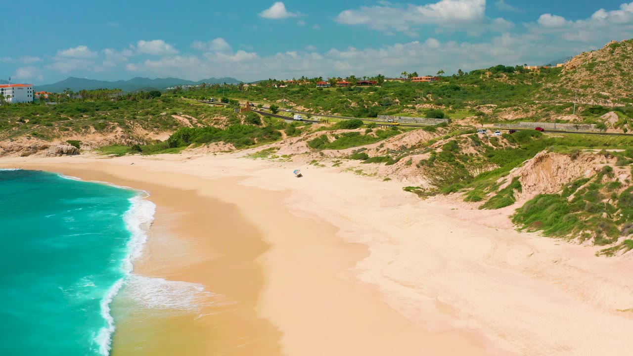 Aerial view of Dreams Beach with a broader strip of turquoise water at left, mild white waves on the long sand, scrub-covered dunes and hills above, road traffic in the distance, and no visible people.