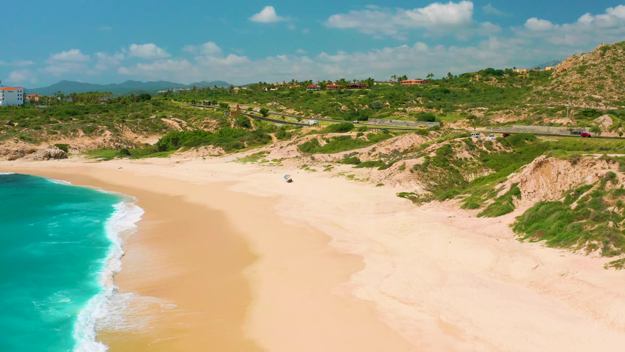 Dreams Beach from above, with the ocean and foamy shore at left, open sand across the lower frame, dune vegetation and eroded slopes in the middle distance, a road with cars, and no visible people.