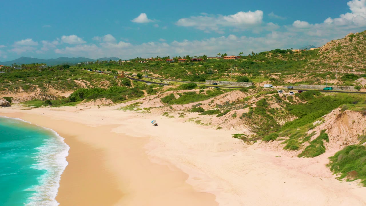 Aerial view at Dreams Beach in Baja California Sur, Mexico, with turquoise surf entering from the left, open pale sand below, green dune scrub, a coastal road, hills, and no visible people.