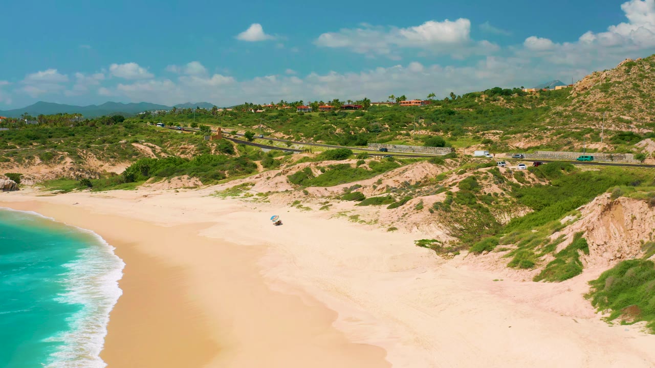 Wide aerial frame of Dreams Beach with the surf strip lower left, broad empty sand, scrub-covered dunes, a road with small vehicles, hillside buildings, and no visible people.
