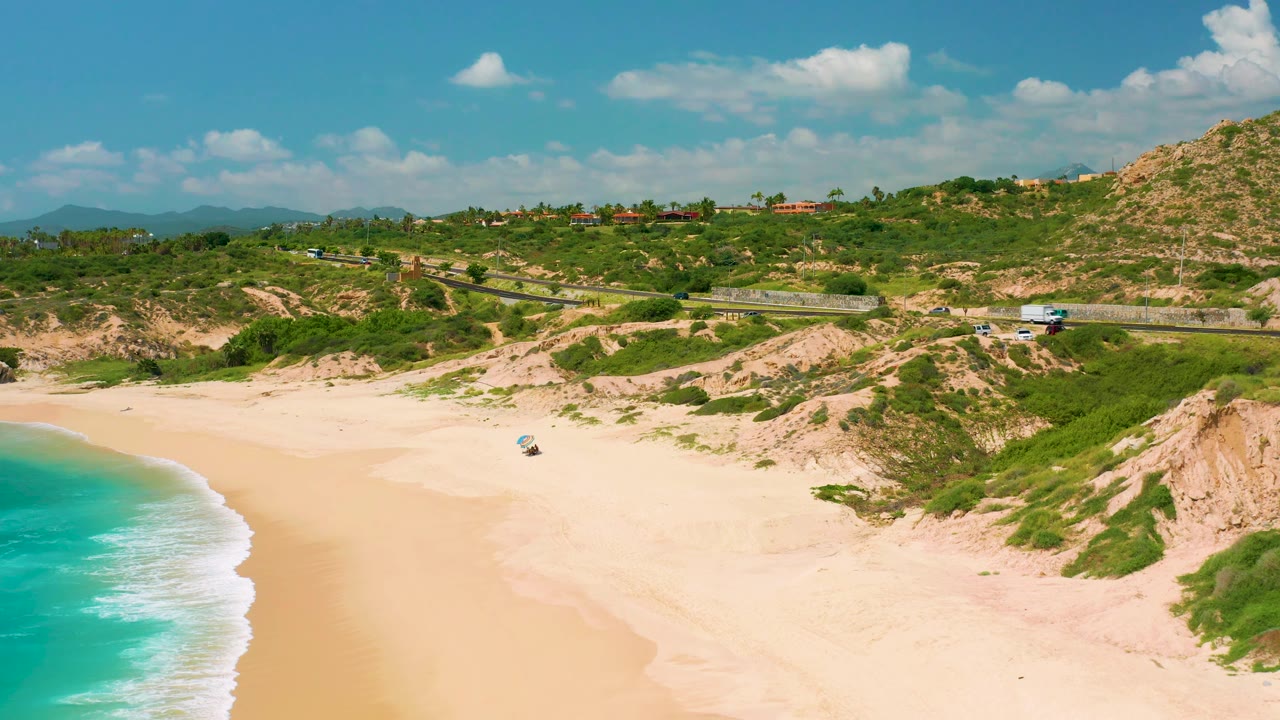Aerial view over Dreams Beach showing aqua water at left, a smooth sand cove, green-and-tan dune ridges, the coastal road, distant rooftops, and no visible people.