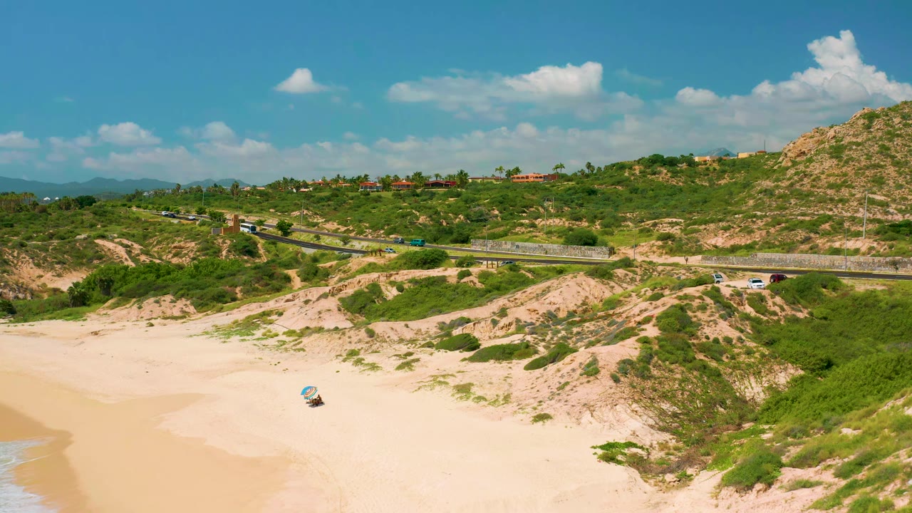 Wide aerial frame at Dreams Beach, Baja California Sur with the road running across scrub-covered dunes, scattered cars on the pavement, rocky hillside at right, and no visible people.