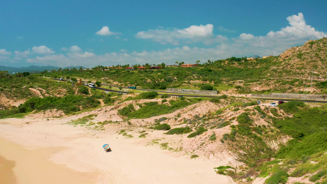 Aerial view at Dreams Beach, Baja California Sur with bright sand below, cars and a bus on the highway, green hillside homes or structures in the distance, and no visible people.