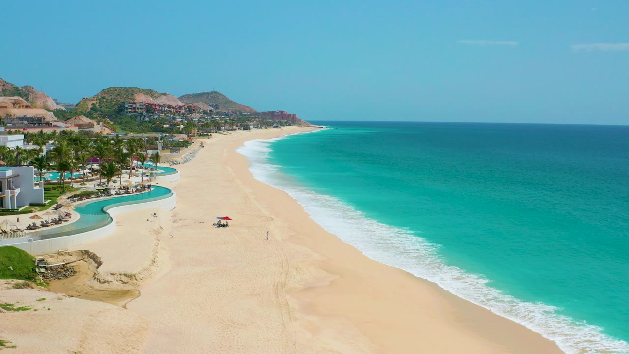 Opening aerial frame over Dreams Beach in Baja California Sur, Mexico, with resort pools and palms at left, a broad sandy beach below, turquoise ocean at right, and tiny distant beach figures.