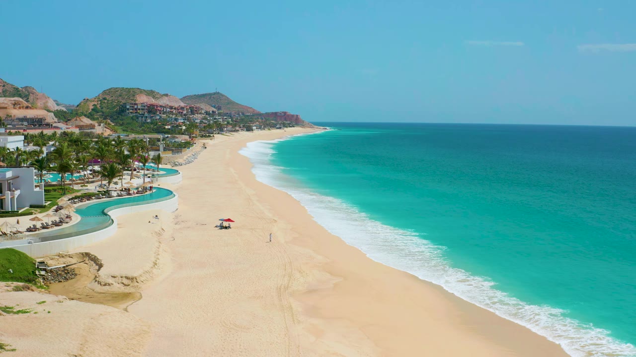 Aerial frame at Dreams Beach showing curved resort pool edges and palms on the left, pale sand through the center, bright turquoise water on the right, and small distant figures near a red umbrella.