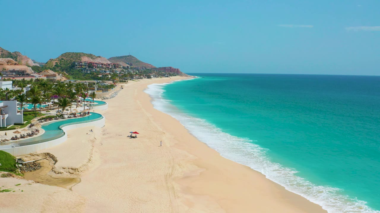Aerial view of Dreams Beach with the resort grounds tight to the left edge, an open sweep of sand through the middle, white waves along the shore, and tiny beach figures under bright sun.