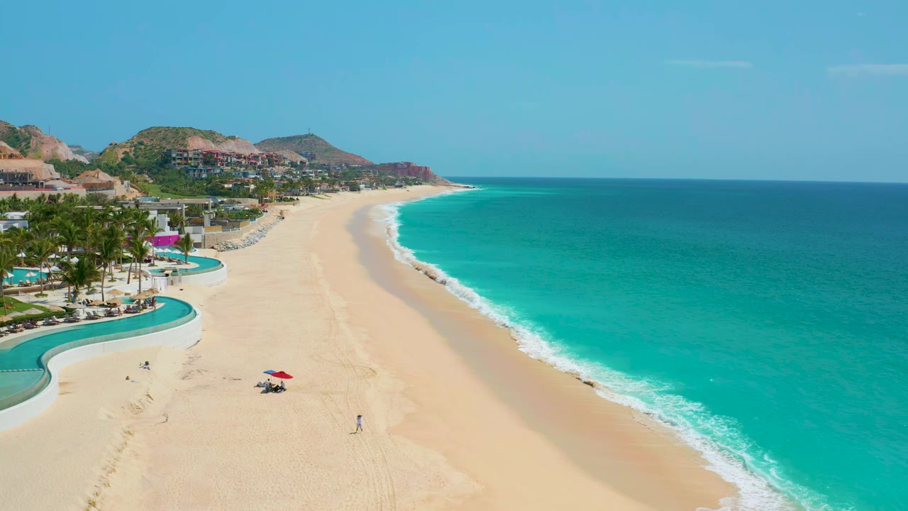 Aerial view along a wide sandy shoreline with turquoise water and resort grounds at left.