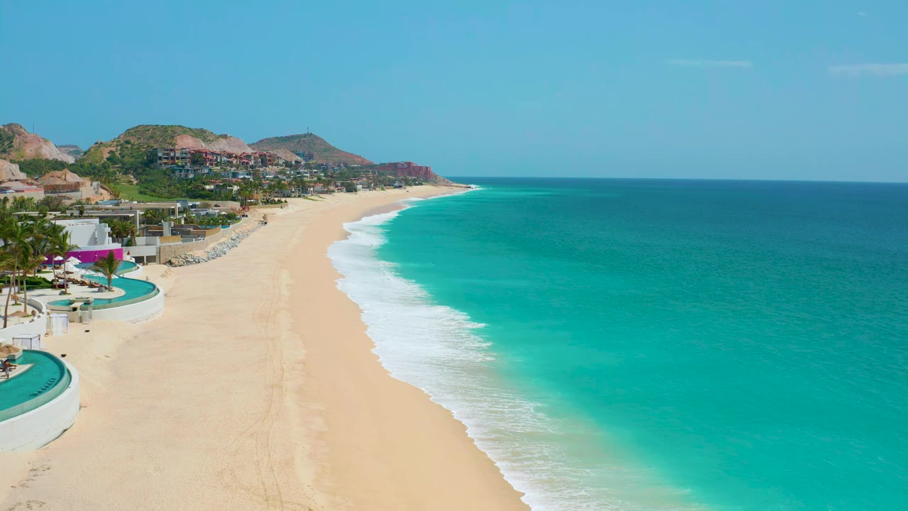 Wide aerial coastline with resort pools, empty sand, surf, and blue water.