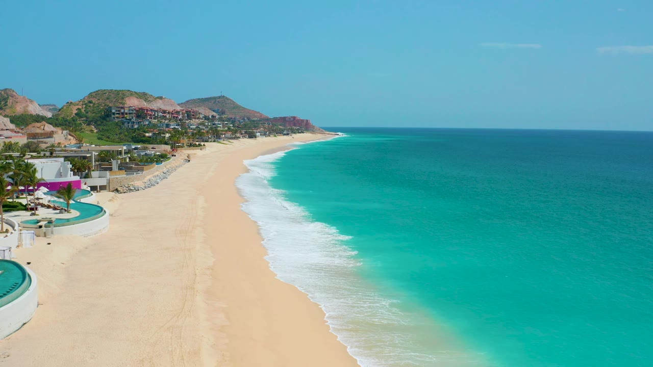 Drone aerial frame from Dreams Beach looking down the coast, with pale sand, a foamy wave band, deepening turquoise water to the right, hillside development beyond the beach, and no visible people.