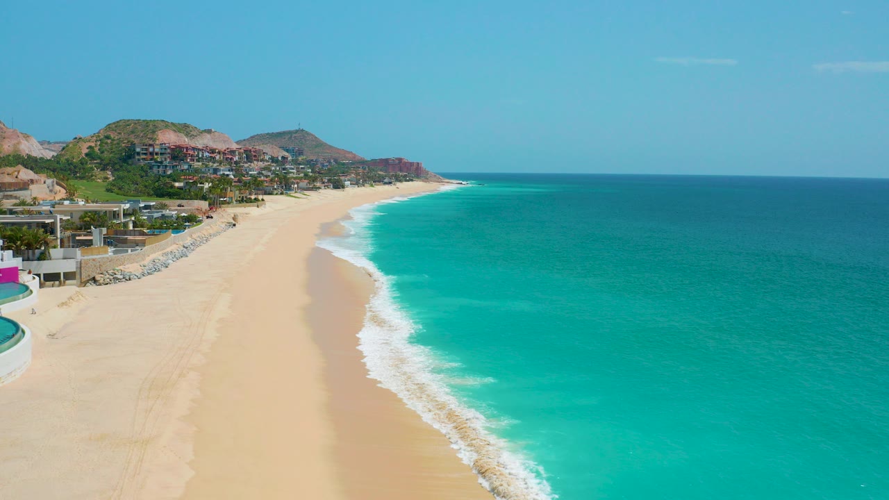 Opening aerial frame of Dreams Beach in Baja California Sur with pale sand, turquoise water, hillside buildings, and no visible people.