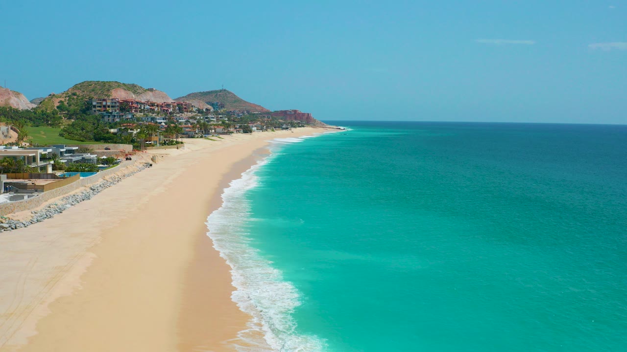 Opening aerial frame over Dreams Beach in Baja California Sur, Mexico, with pale sand at left, turquoise surf at right, hillside buildings in the distance, and no visible people.
