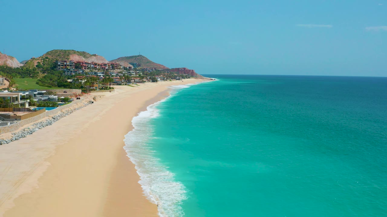 Aerial frame from Dreams Beach in Baja California Sur, Mexico, showing a broad turquoise sea, a narrow white surf band on the sand, hillside development at left, and no visible people.