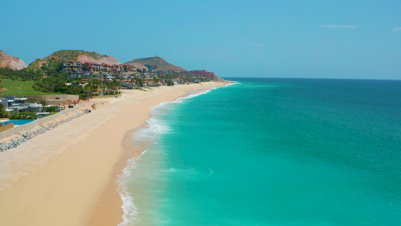 Opening aerial view at Dreams Beach in Baja California Sur, Mexico, showing a curving sandy beach, turquoise ocean, hillside homes, clear sky, and no visible people.