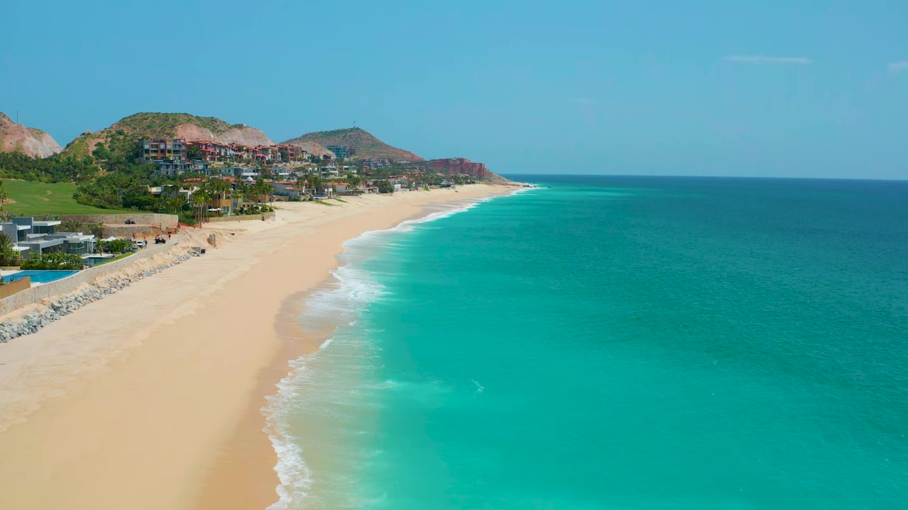Aerial frame at 0:01 over Dreams Beach, Baja California Sur, Mexico, with white surf tracing the sand, blue-green water filling the right side, hillside homes beyond, and no visible people.