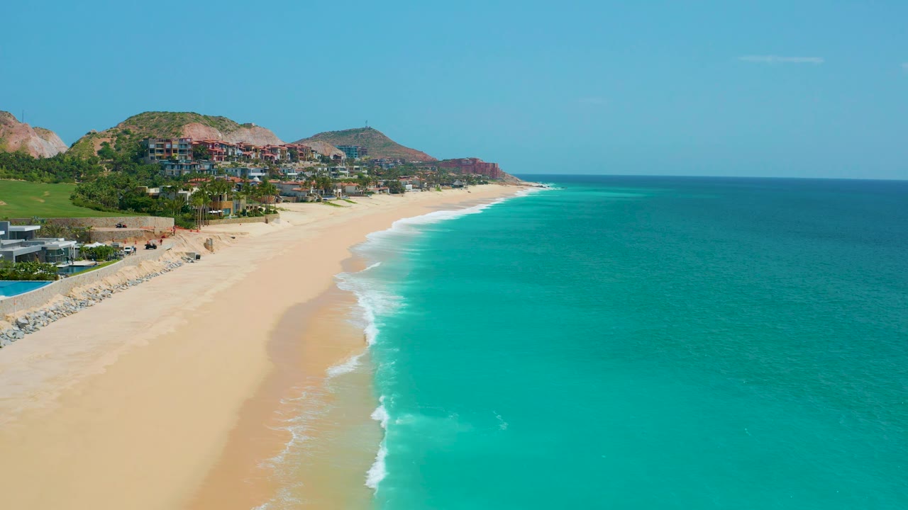 Elevated beach view with pale sand, surf, ocean, and hillside residences.