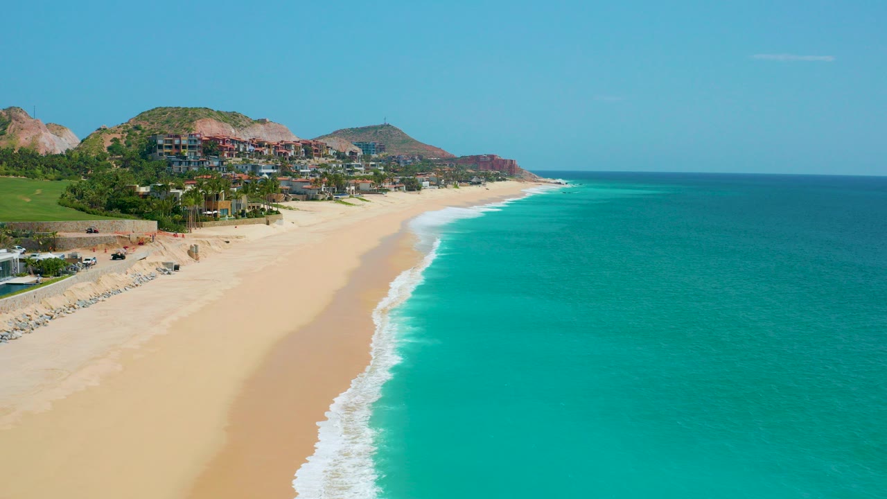 Opening aerial view at Dreams Beach, Baja California Sur, Mexico, showing turquoise water, a wide sandy shore, distant hillside buildings, and no visible people.