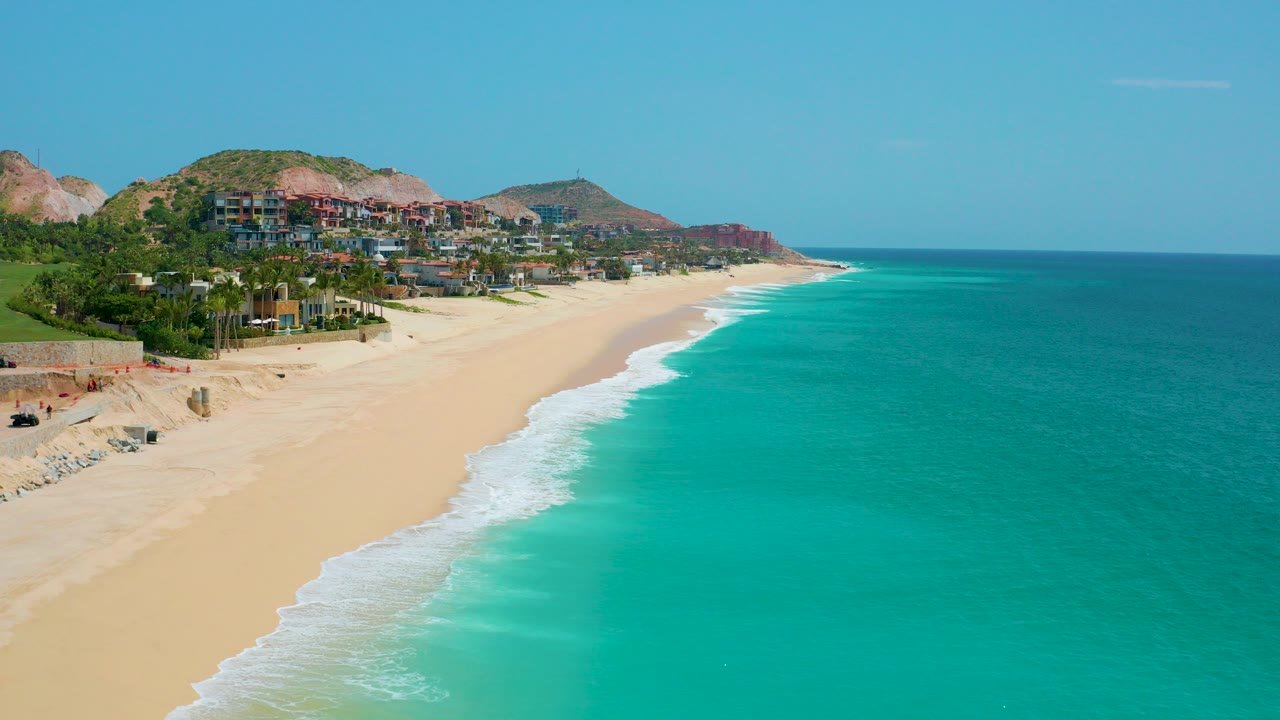 Aerial view over Dreams Beach in Baja California Sur, Mexico, with a wide sandy shoreline, teal water, hillside homes, and no visible people at the start of the clip.