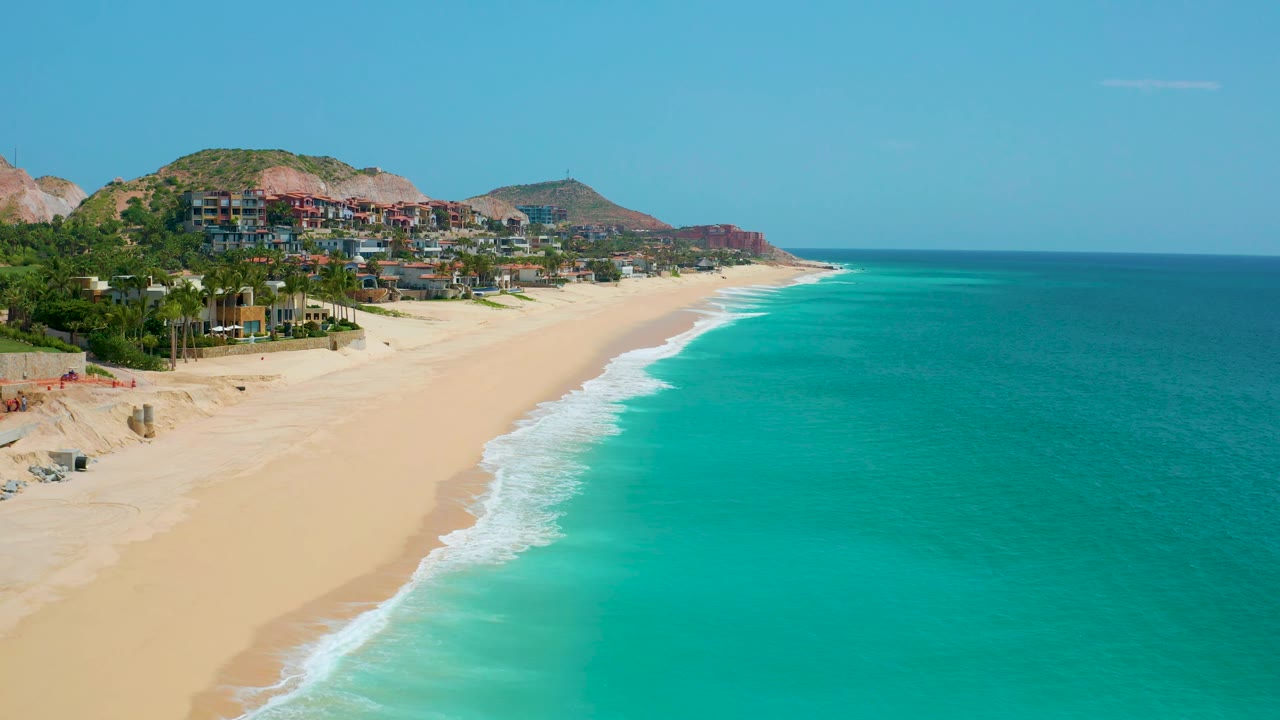 Aerial beach view with foam patterns, teal water, and hillside homes.