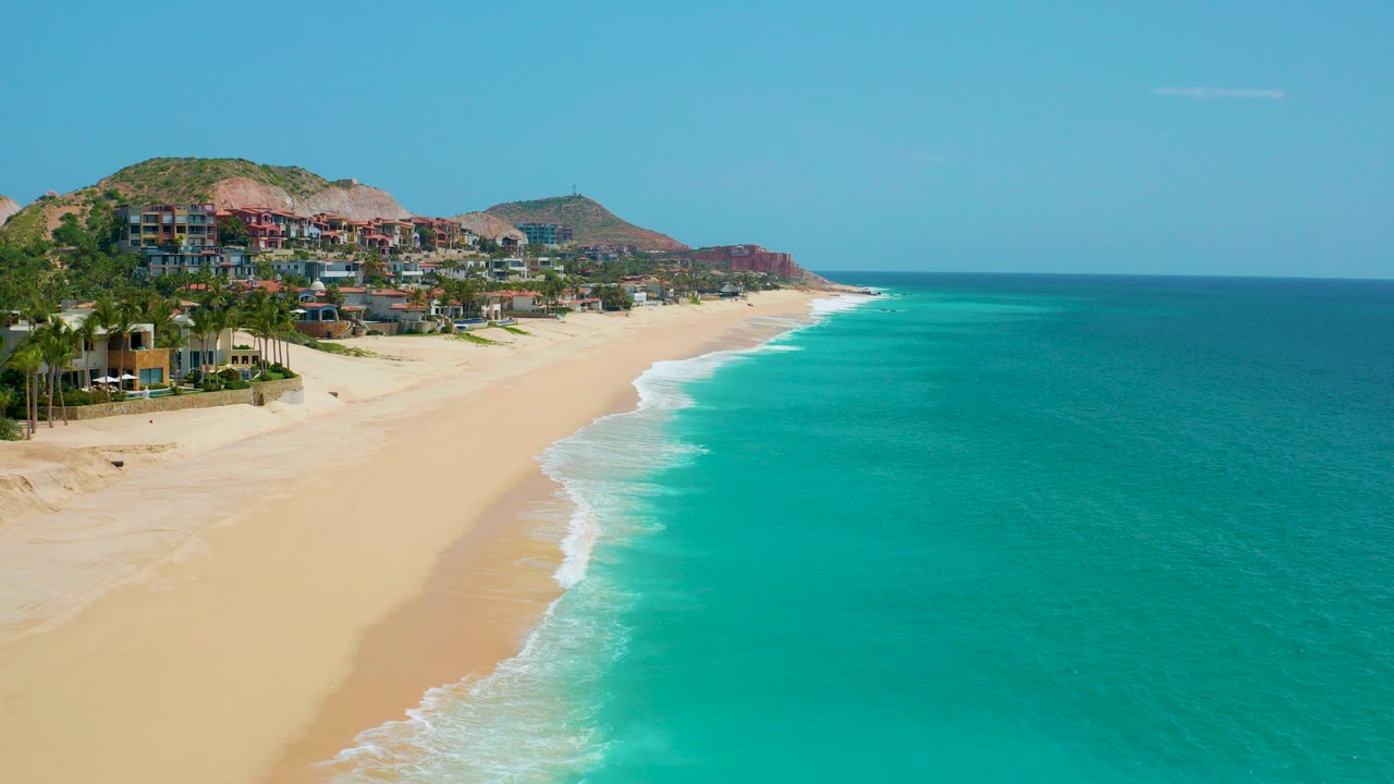Aerial frame over Dreams Beach in Baja California Sur showing pale sand, turquoise water, hillside buildings, clear sky, and no visible people at the start of the clip.