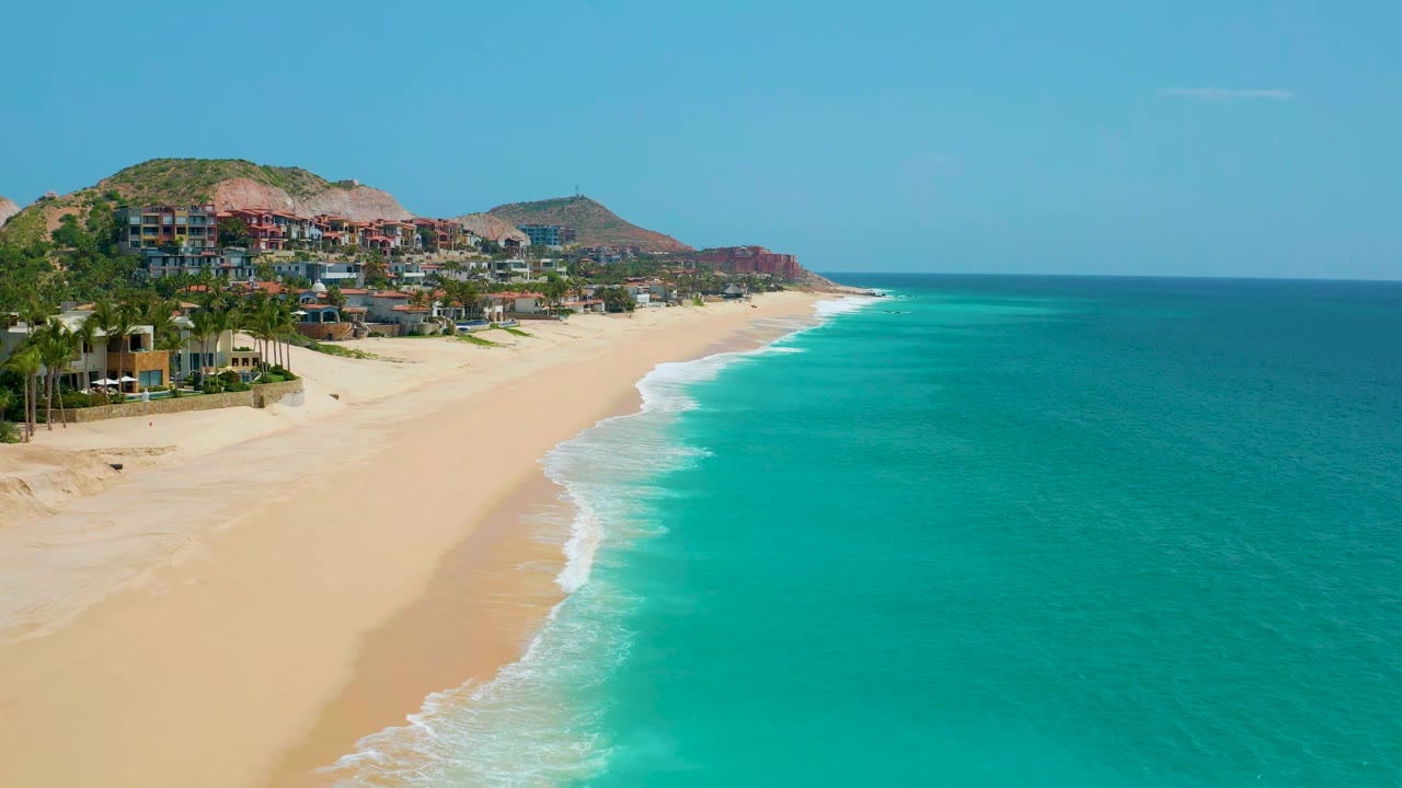 A wide aerial view of Dreams Beach shows the shoreline bending away, white waves tracing the sand, hillside structures behind the beach, and no visible people.