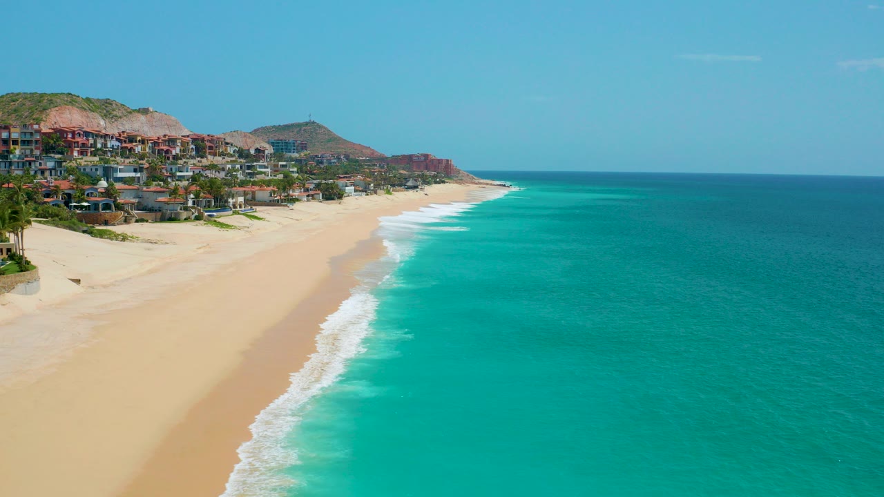 Opening frame of Dreams Beach in Baja California Sur, Mexico, with turquoise water on the right, pale sand on the left, hillside buildings in the distance, and no visible people.