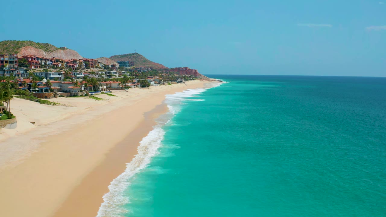 Wide beach coastline with turquoise water, white shore break, and buildings along the hills.