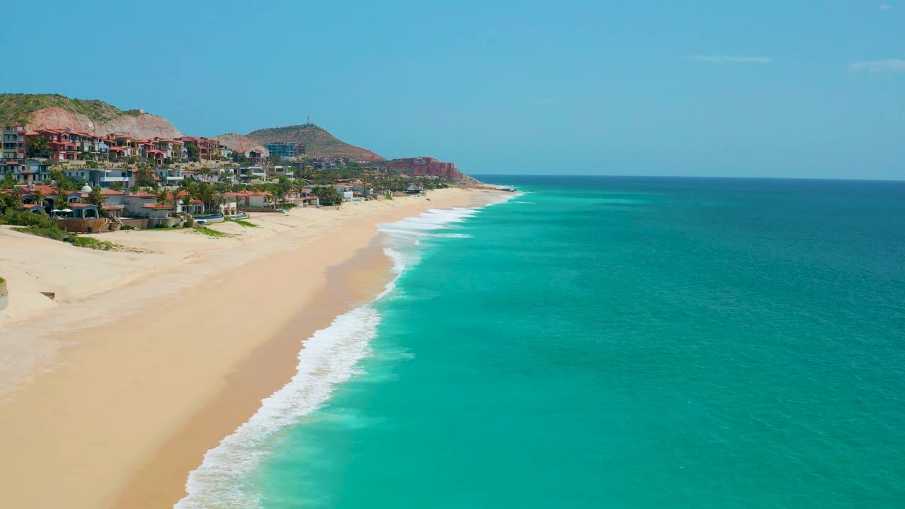 At 0:02, the diagonal shoreline of Dreams Beach divides pale sand from bright turquoise ocean, with hillside development and low hills in the background and no visible people.