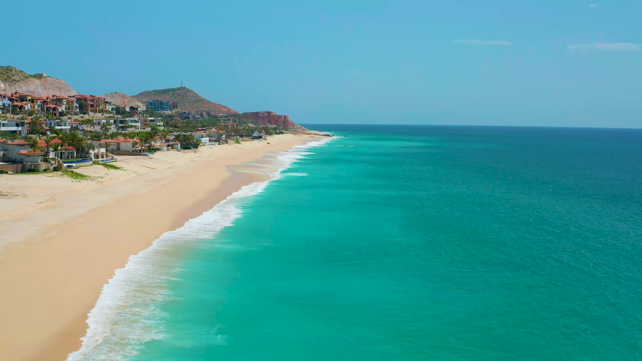 Wide coastal view with surf line, open blue-green water, beach homes, and hills.