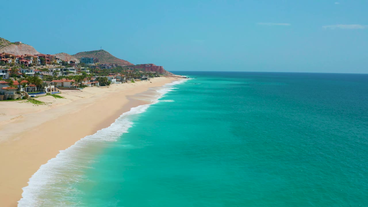 Aerial coastal frame at Dreams Beach with more open turquoise water filling the right side, pale sand and foam at left, distant homes along the shore, and no visible people.