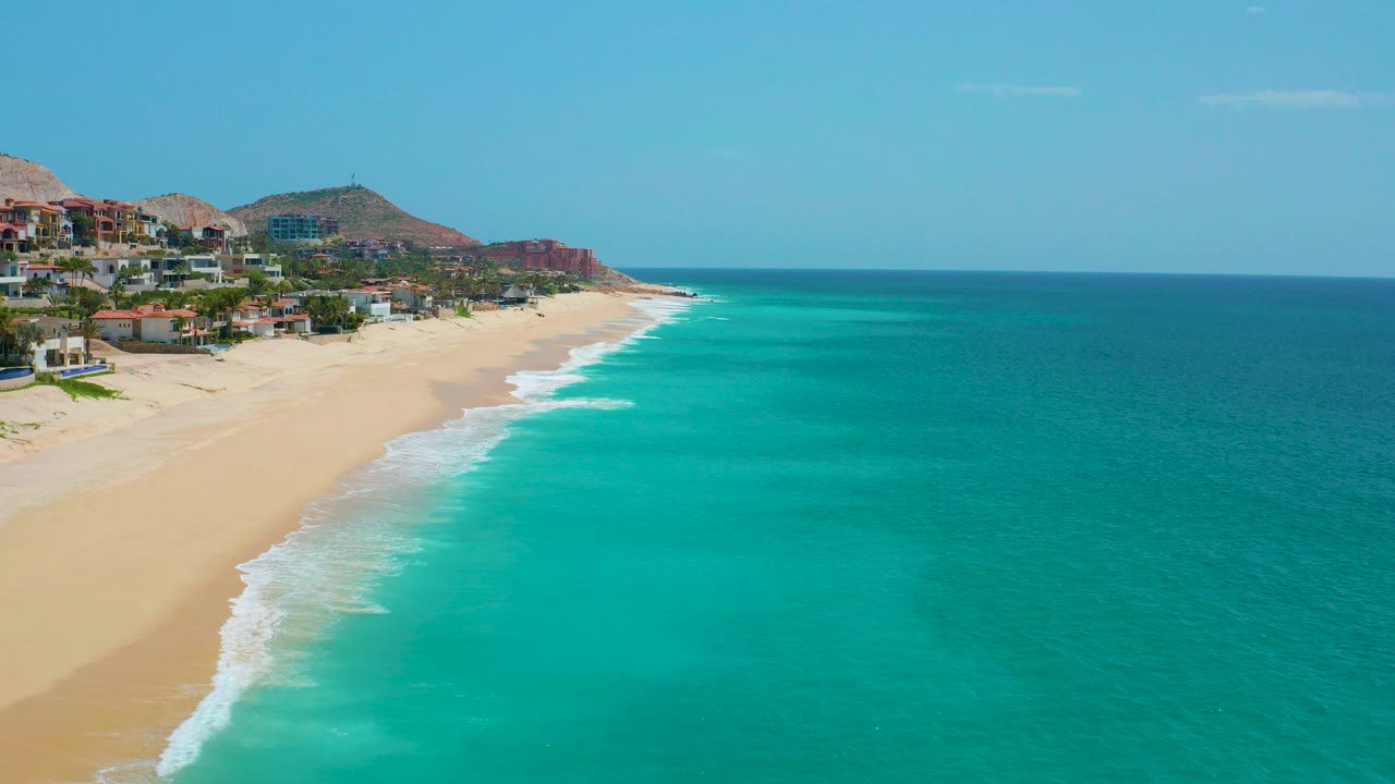 Opening aerial frame at Dreams Beach, Baja California Sur, with turquoise surf meeting a wide sandy shoreline, hillside houses on the left, and no visible people.