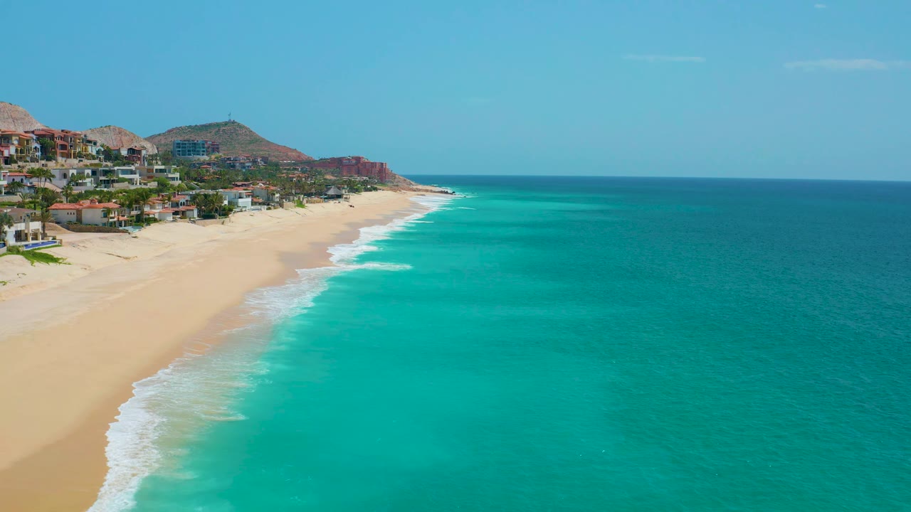 Aerial view over Dreams Beach with more open ocean filling the right side, surf breaking in thin white bands, hillside homes at left, and no visible people.