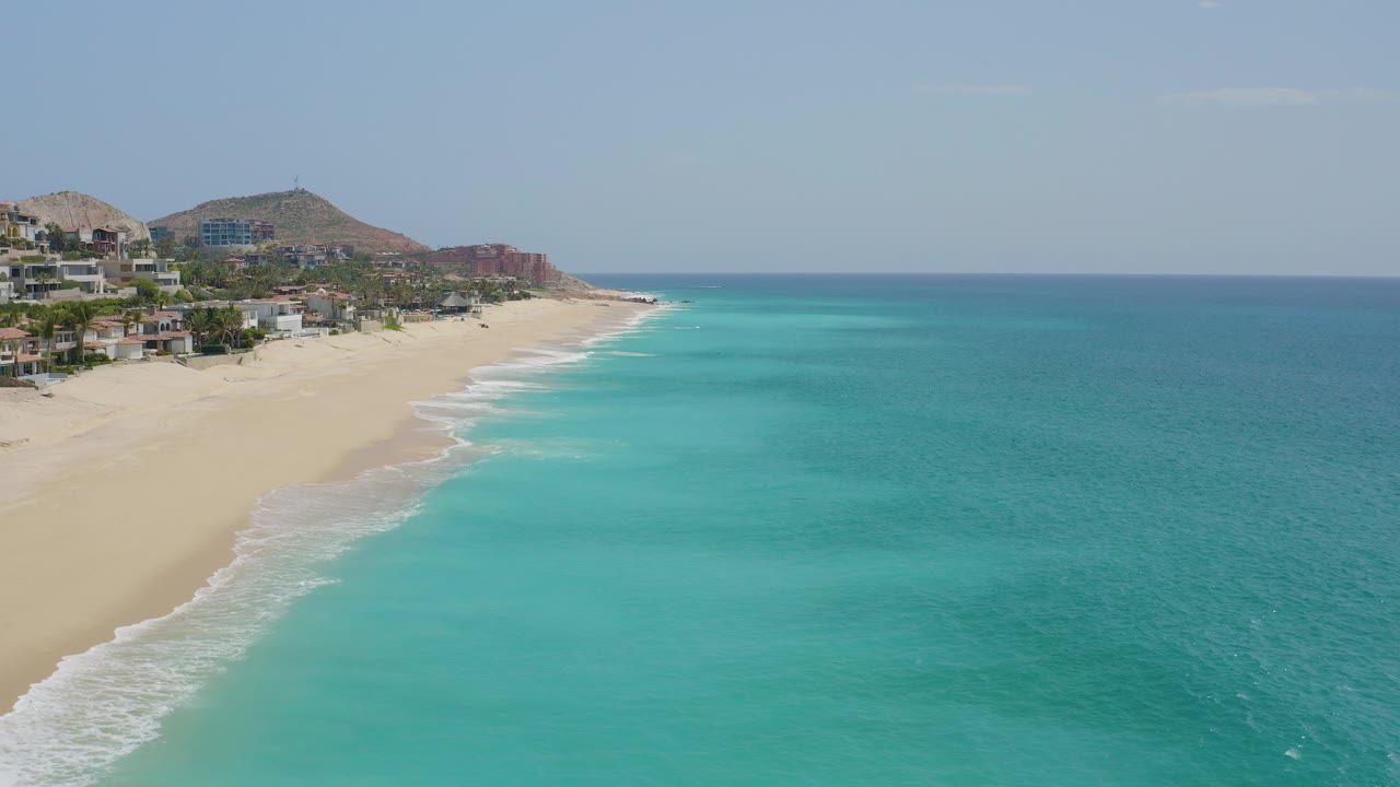 Aerial frame over Dreams Beach in Baja California Sur with pale sand at left, turquoise shallows at right, hillside buildings in the background, and no visible people.