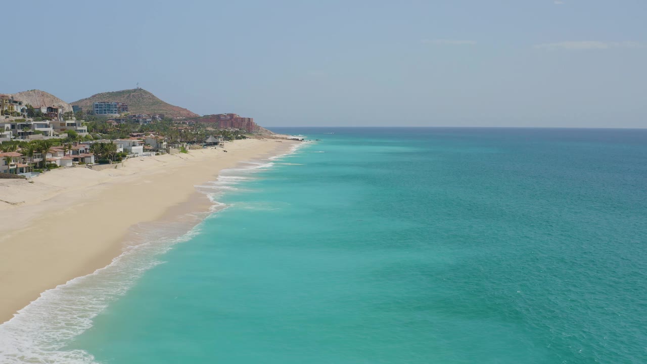 Dreams Beach appears as a broad strip of sand with white wave wash, turquoise shallows, darker offshore water, hillside structures, and no visible people.