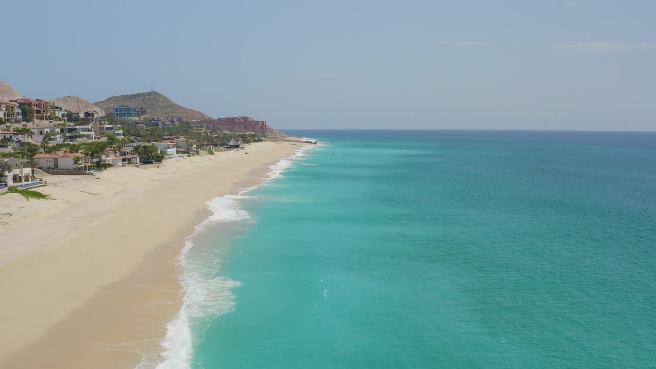 Aerial view of Dreams Beach in Baja California Sur with pale sand, turquoise water, white surf along the shore, hillside buildings in the distance, and no visible people.