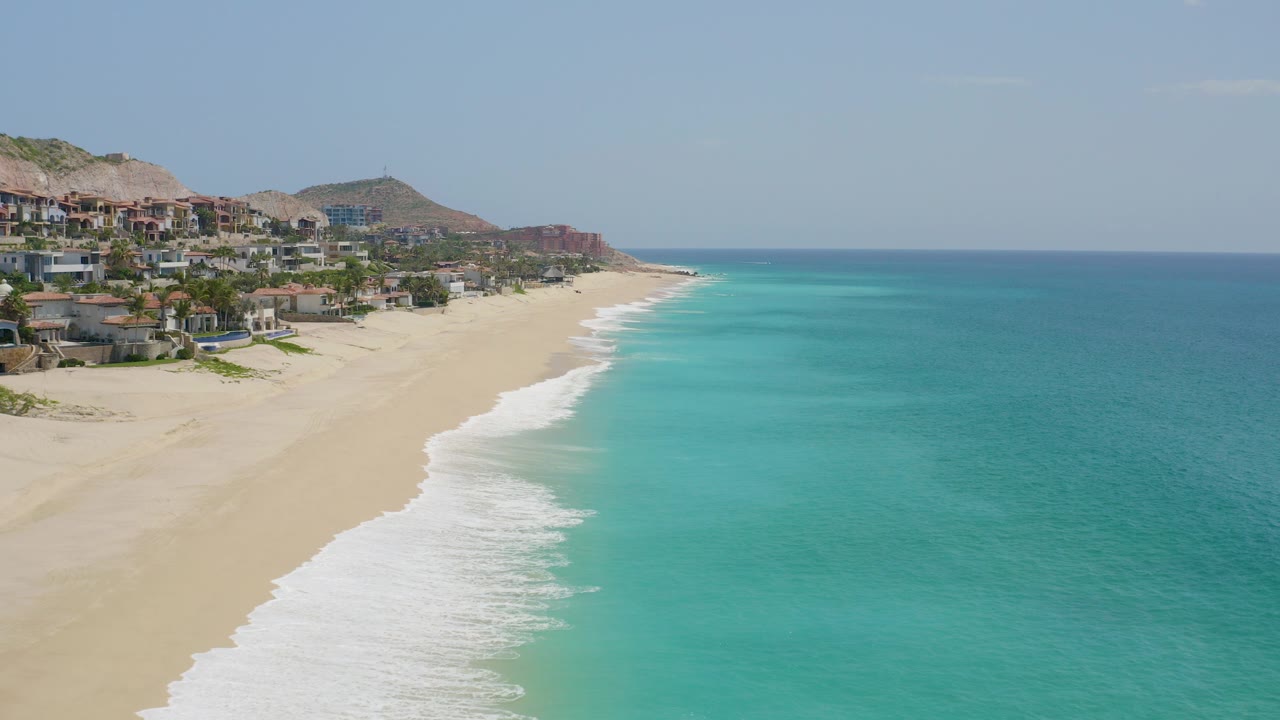 Opening frame over Dreams Beach in Baja California Sur, Mexico, showing hillside oceanfront homes at left, turquoise water at right, a pale horizon, and no visible people.