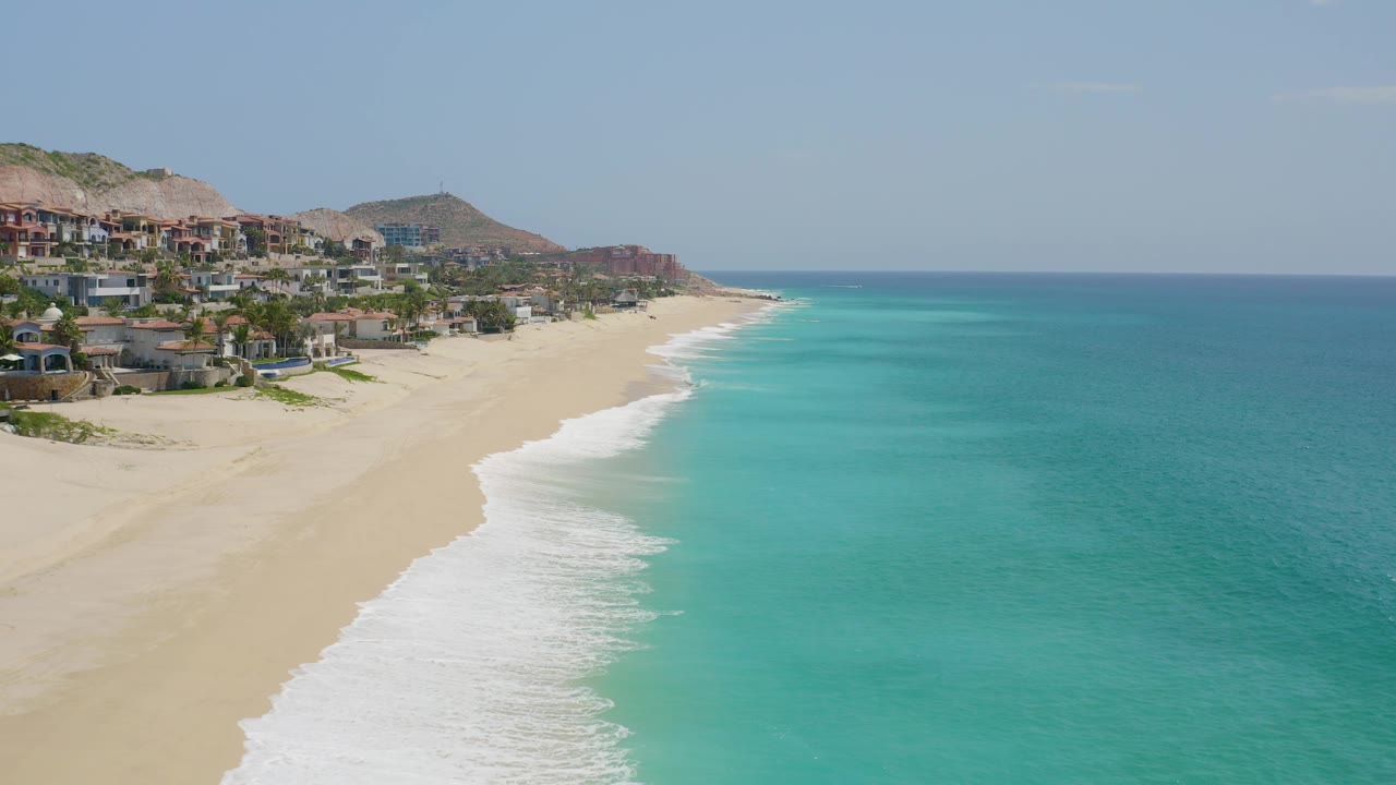 Third frame at Dreams Beach in Baja California Sur, Mexico, showing more of the beachfront homes and palms on the left edge, foamy surf across the sand, turquoise sea, and no visible people.