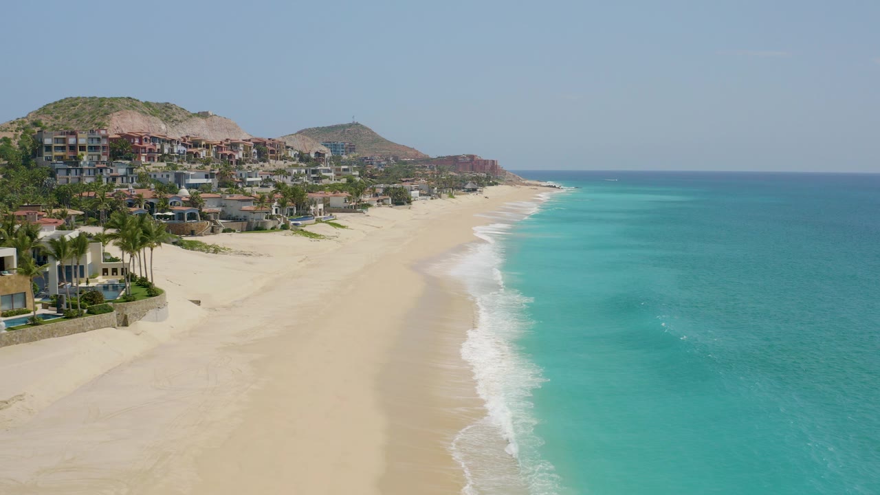 Wide aerial frame at Dreams Beach, Baja California Sur, Mexico showing a long sandy shoreline, turquoise ocean to the right, hillside resort buildings to the left, and no visible people.