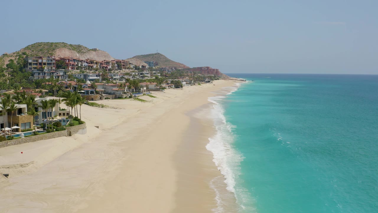 Dreams Beach coastline frame showing blue-green water on the right, a sweeping sand beach, hillside buildings in the distance, dry hills behind them, and no visible people.