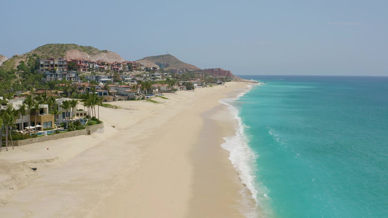 Aerial beach view with palms, resort structures, pale sand, and ocean surf.