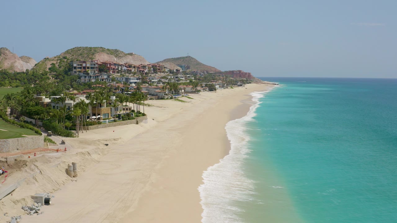 Aerial view at Dreams Beach in Baja California Sur showing pale sand, turquoise water, hillside buildings, and a white surf edge with no visible people.