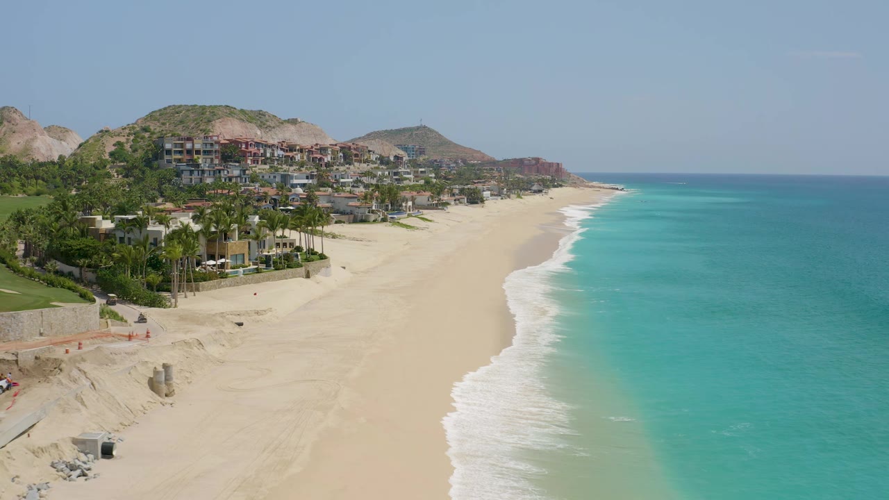 Wide beach shoreline with blue water, surf, landscaped coast, and hillside buildings