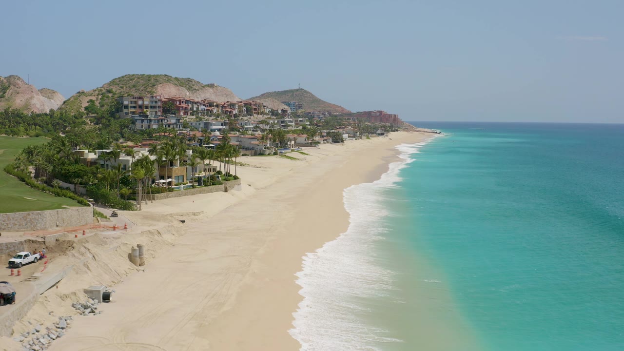 Aerial view of Dreams Beach with a curving surf line, turquoise water, hillside structures, small vehicles near the lower beach edge, and no visible people.