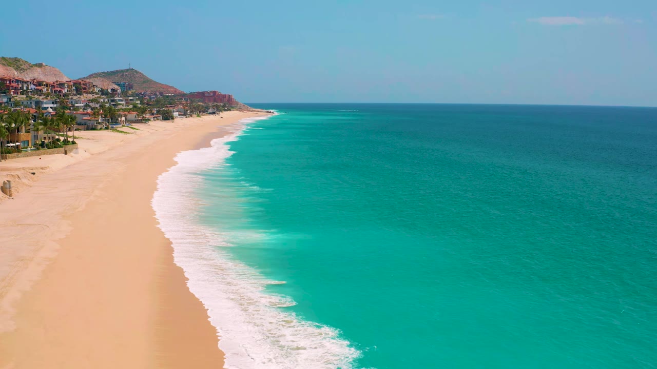 Curving beach and turquoise water with white waves and distant hillside buildings.