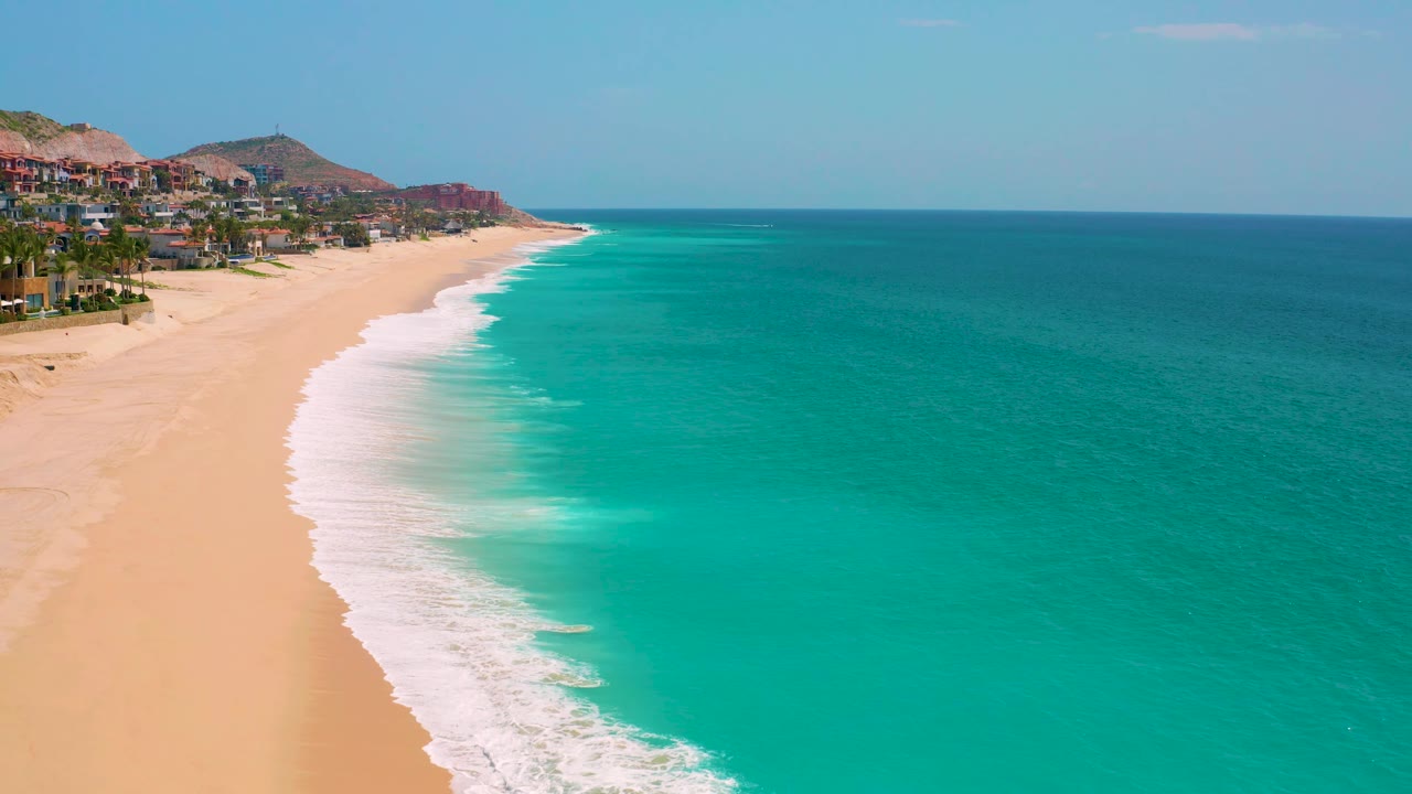 Aerial frame over Dreams Beach in Baja California Sur, Mexico, showing more open water across the right side, pale sand along the left, foamy surf, distant buildings, and no visible people.