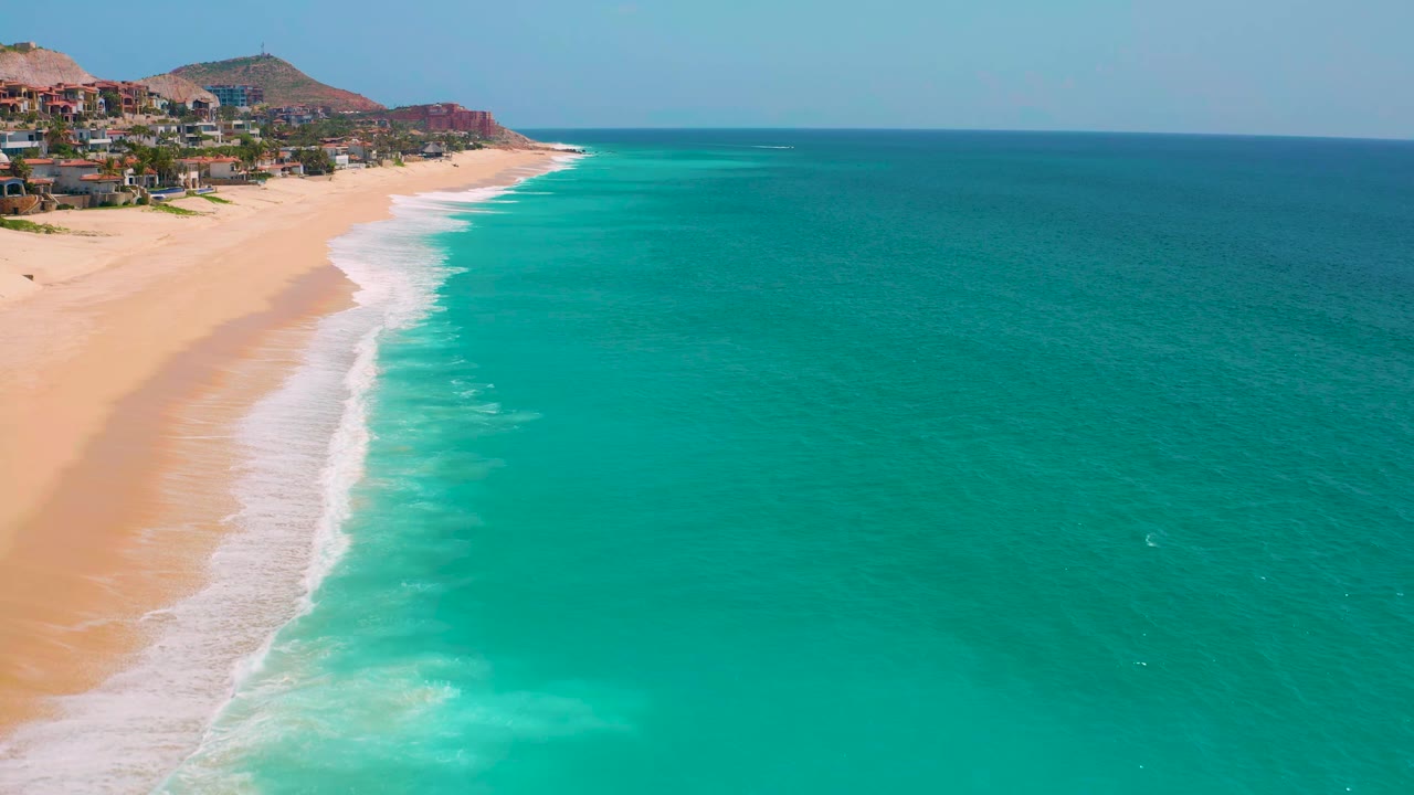 Curving beach and surf line beside broad turquoise water with buildings near low hills.